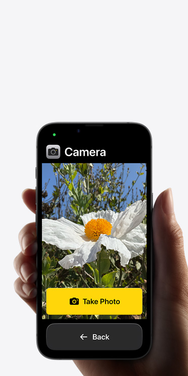 iPhone 16e screen showing simplified Camera app interface with a white flower in the frame, “Take Photo” button and “Back” button below