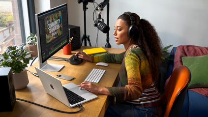 A woman in her home working at her desk with MacBook Pro connected to an external display, wearing headphones and speaking into a microphone